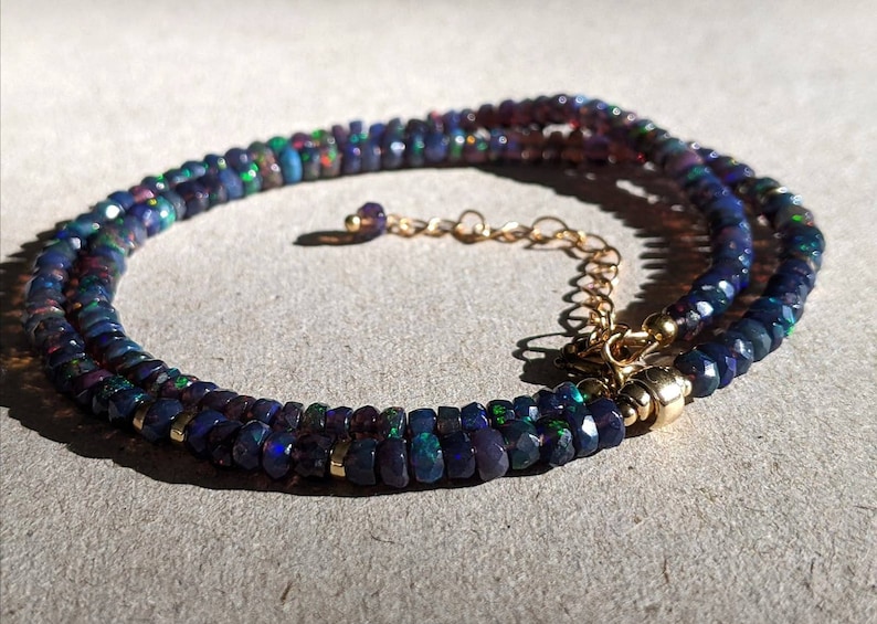 Op de afbeelding: Een ketting met gefacetteerde donkerblauwe en zwarte opaal kralen met iriserende groene en rode flitsen. De streng is geaccentueerd met kleine goudkleurige metalen tussenstukjes en bevat een goudkleurige metalen verlengketting met karabijnsluiting.