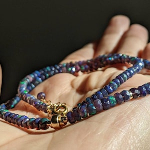 Op de afbeelding: Een ketting bestaande uit gefacetteerde, donkerblauwe opaal kralen met iriserende flitsen van groen en rood. Het snoer is bevestigd met een gepolijste goudkleurige metalen sluiting en een kleine ronde ring, wat een glinsterend, cirkelvormig ontwerp vormt.