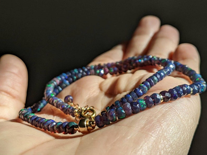 Op de afbeelding: Een ketting bestaande uit gefacetteerde, donkerblauwe opaal kralen met iriserende flitsen van groen en rood. Het snoer is bevestigd met een gepolijste goudkleurige metalen sluiting en een kleine ronde ring, wat een glinsterend, cirkelvormig ontwerp vormt.