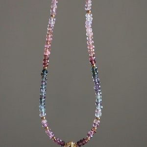 May include: A necklace with a gradient of gemstones in shades of pink, purple, blue, and green. Small gold-coloured beads are interspersed throughout. The necklace has a gold magnetic clasp. A stylish piece of jewellery.