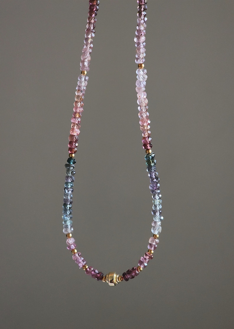 May include: A necklace with a gradient of gemstones in shades of pink, purple, blue, and green. Small gold-coloured beads are interspersed throughout. The necklace has a gold magnetic clasp. A stylish piece of jewellery.