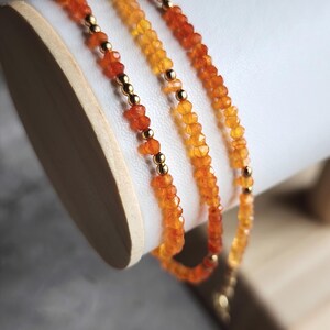 May include: Three strands of orange gemstone bead necklaces with gold-colored spacer beads. The necklaces are draped over a white cylindrical display stand. The gemstones vary in color from light yellow to deep orange.