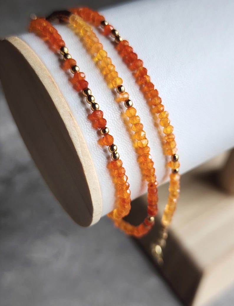 May include: Three strands of orange gemstone bead necklaces with gold-colored spacer beads. The necklaces are draped over a white cylindrical display stand. The gemstones vary in color from light yellow to deep orange.