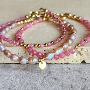 Puede incluir: Tres pulseras delicadas con piedras preciosas rosas, perlas brillantes y cuentas doradas. Una pulsera tiene un peque&ntilde;o dije redondo dorado. Las pulseras est&aacute;n apiladas, creando un look de joyer&iacute;a en capas.