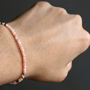 May include: A delicate bracelet made of small pink and white beads with gold accents. The bracelet is worn on a person's wrist.