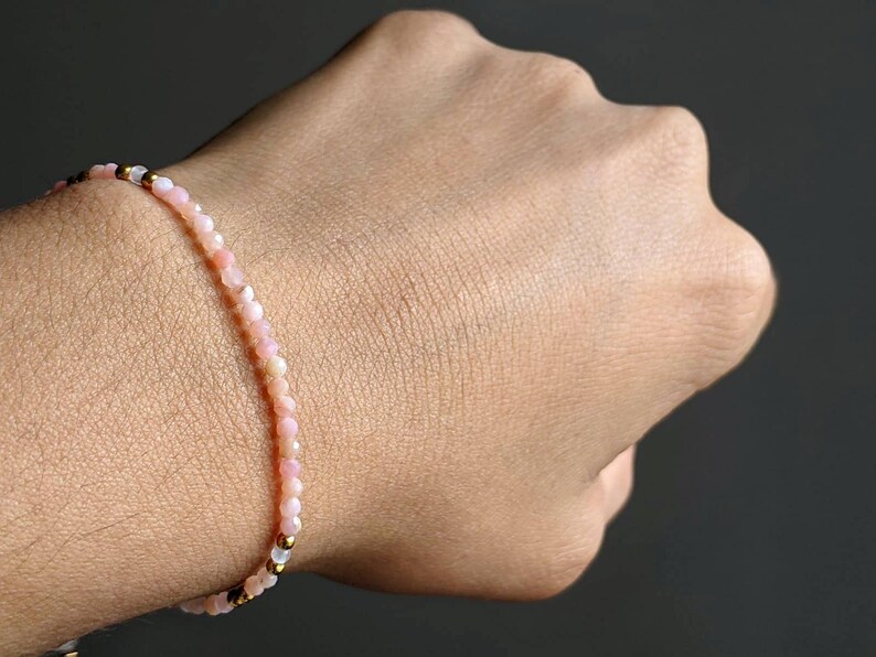 May include: A delicate bracelet made of small pink and white beads with gold accents. The bracelet is worn on a person's wrist.