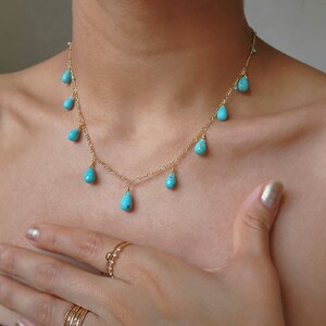 May include: A delicate gold necklace featuring turquoise teardrop-shaped pendants. The necklace is displayed on a person's neck and chest, with a hand and rings visible in the frame. The turquoise stones are a vibrant blue-green color.