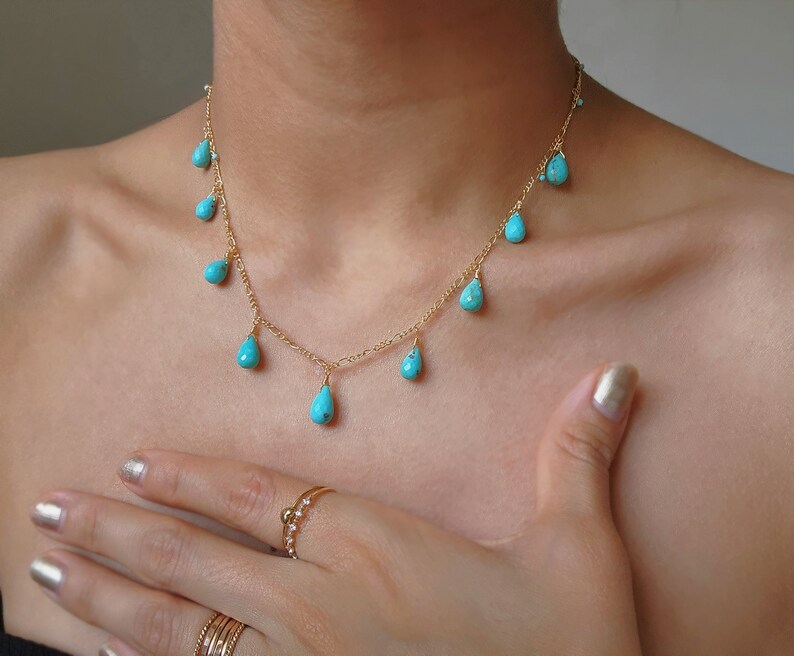 May include: A delicate gold necklace featuring turquoise teardrop-shaped pendants. The necklace is displayed on a person's neck and chest, with a hand and rings visible in the frame. The turquoise stones are a vibrant blue-green color.