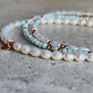 May include: Close-up of three delicate pearl bracelets. The bracelets feature white pearls, small blue-green beads, and rose gold accents. The pearls have a lustrous sheen, and the bracelets are arranged on a textured surface.