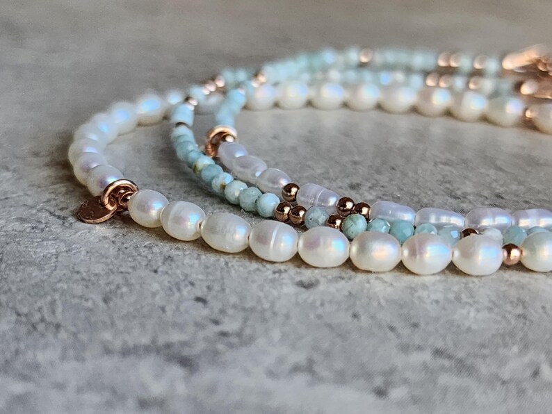 May include: Close-up of three delicate pearl bracelets. The bracelets feature white pearls, small blue-green beads, and rose gold accents. The pearls have a lustrous sheen, and the bracelets are arranged on a textured surface.
