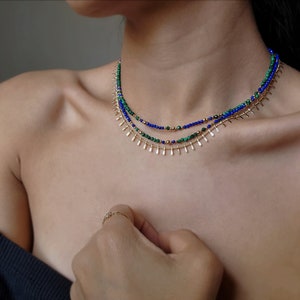 May include: A layered necklace with green and blue beads and gold accents. The necklace is made of small, round beads and has a delicate, feminine look.