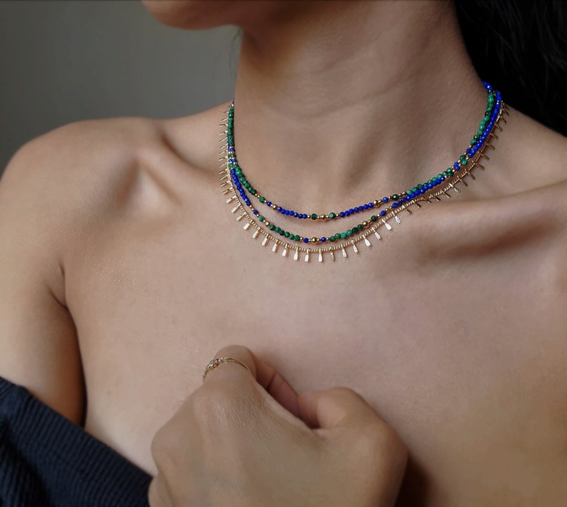 May include: A layered necklace with green and blue beads and gold accents. The necklace is made of small, round beads and has a delicate, feminine look.