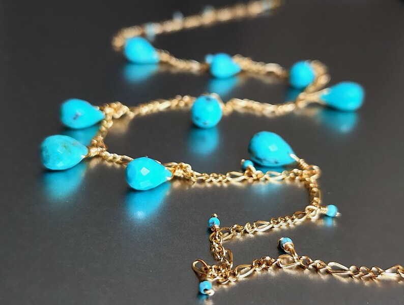 May include: A delicate gold necklace featuring teardrop-shaped turquoise beads. The necklace has a fine chain and is displayed on a reflective surface. The turquoise beads are a vibrant blue-green color.