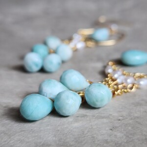 May include: A pair of gold-toned earrings featuring light blue, teardrop-shaped gemstones and small white pearls. The earrings have a delicate design with multiple gemstones and pearls, creating an elegant look.