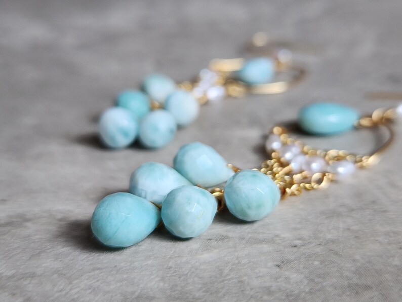 May include: A pair of gold-toned earrings featuring light blue, teardrop-shaped gemstones and small white pearls. The earrings have a delicate design with multiple gemstones and pearls, creating an elegant look.