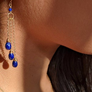 May include: Gold-toned earrings with three dangling, teardrop-shaped, deep blue gemstones. The earrings feature a delicate chain design and a small blue bead at the top. The jewelry is shown against a blurred background.