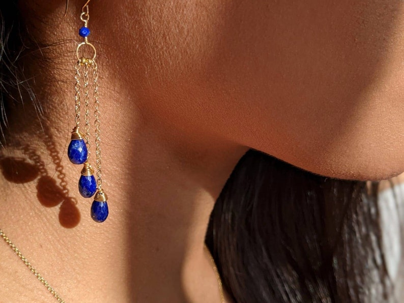May include: Gold-toned earrings with three dangling, teardrop-shaped, deep blue gemstones. The earrings feature a delicate chain design and a small blue bead at the top. The jewelry is shown against a blurred background.
