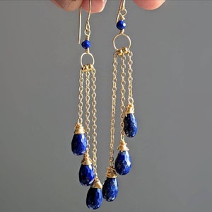 May include: Gold-tone dangle earrings featuring faceted, teardrop-shaped, deep blue lapis lazuli gemstones. The earrings have delicate gold chains and small blue beads at the top, creating an elegant and sophisticated look.