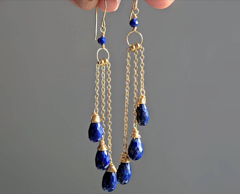 May include: Gold-tone dangle earrings featuring faceted, teardrop-shaped, deep blue lapis lazuli gemstones. The earrings have delicate gold chains and small blue beads at the top, creating an elegant and sophisticated look.
