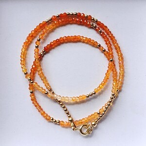 May include: A layered necklace featuring small, faceted orange and yellow beads interspersed with gold-colored beads. The necklace has a gold-colored clasp and is displayed against a white background. The beads create a gradient effect, transitioning from lighter to darker shades.