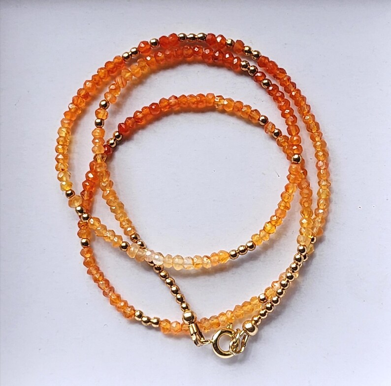 May include: A layered necklace featuring small, faceted orange and yellow beads interspersed with gold-colored beads. The necklace has a gold-colored clasp and is displayed against a white background. The beads create a gradient effect, transitioning from lighter to darker shades.