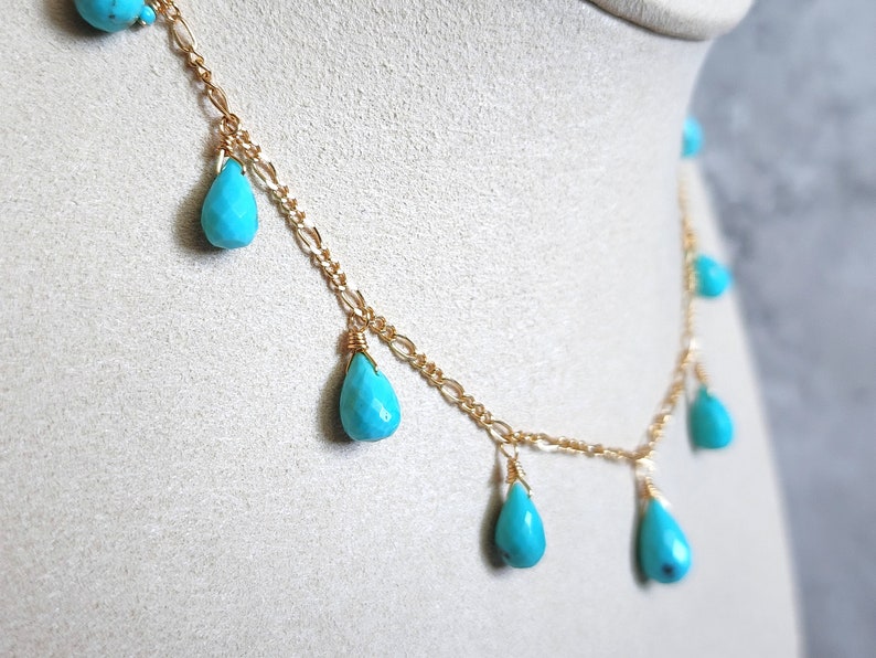 May include: A delicate gold chain necklace featuring teardrop-shaped turquoise beads. The necklace has a delicate chain design with multiple turquoise pendants, creating a layered effect. The turquoise beads are a vibrant blue-green color.