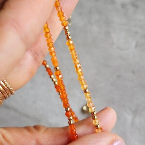 May include: Close-up of a delicate necklace featuring small, faceted orange and yellow beads. The beads are interspersed with small gold-colored beads. The necklace is held up against a gray background.