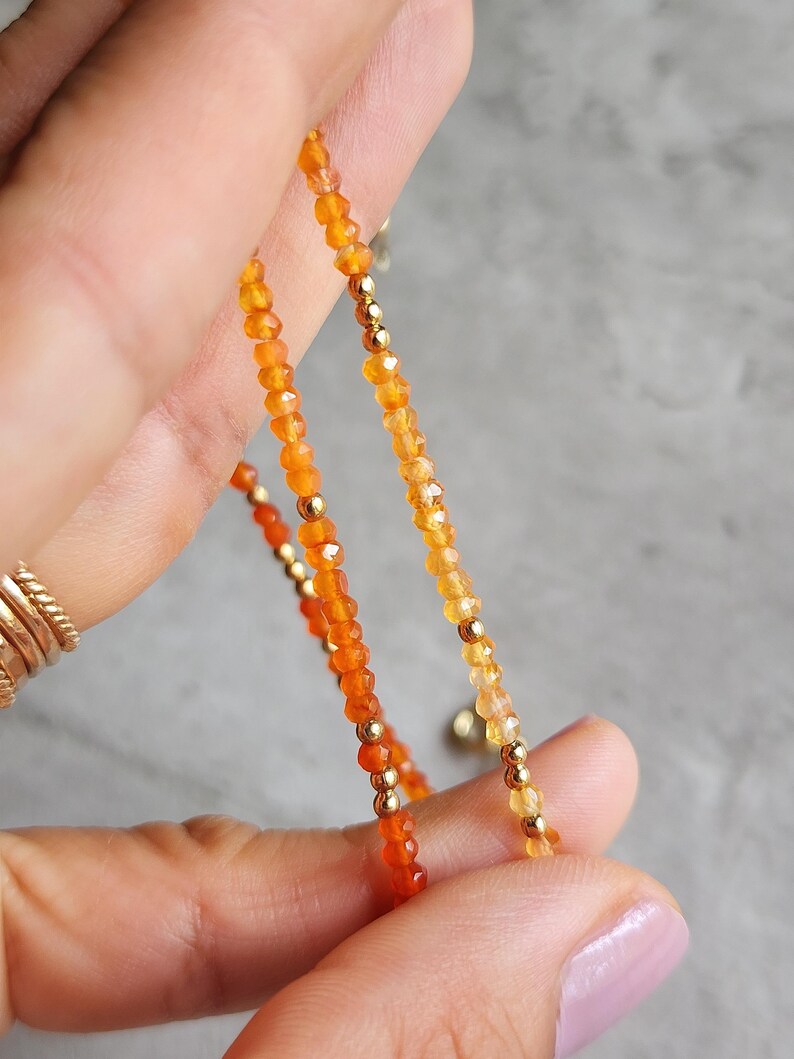 May include: Close-up of a delicate necklace featuring small, faceted orange and yellow beads. The beads are interspersed with small gold-colored beads. The necklace is held up against a gray background.