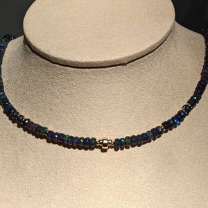 Op de afbeelding: Een ketting bestaande uit een doorlopende streng van kleine, gefacetteerde donkerblauwe en zwarte kralen met iriserende groene en rode vlekjes. De kralen zijn verbonden met een enkele, gepolijste goudkleurige cilindrische kraal in het midden.