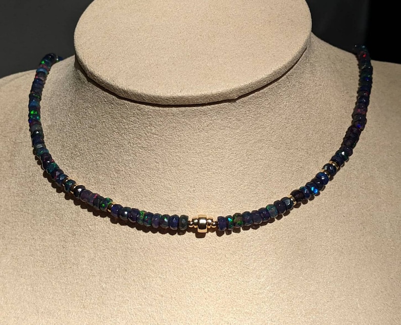 Op de afbeelding: Een ketting bestaande uit een doorlopende streng van kleine, gefacetteerde donkerblauwe en zwarte kralen met iriserende groene en rode vlekjes. De kralen zijn verbonden met een enkele, gepolijste goudkleurige cilindrische kraal in het midden.