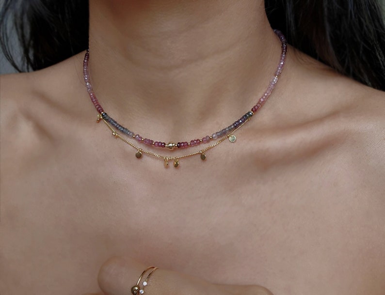 May include: A necklace with a strand of small, multi-coloured beads in shades of pink, purple, and gold. A second, gold chain hangs below, adorned with small, dangling gold discs. The jewelry is displayed on a person's skin.