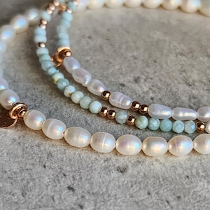May include: Three layered bracelets with white oval pearls, small light blue beads, and rose gold accents. The bracelets are arranged in a curved pattern, showcasing the different textures and colors of the jewelry.