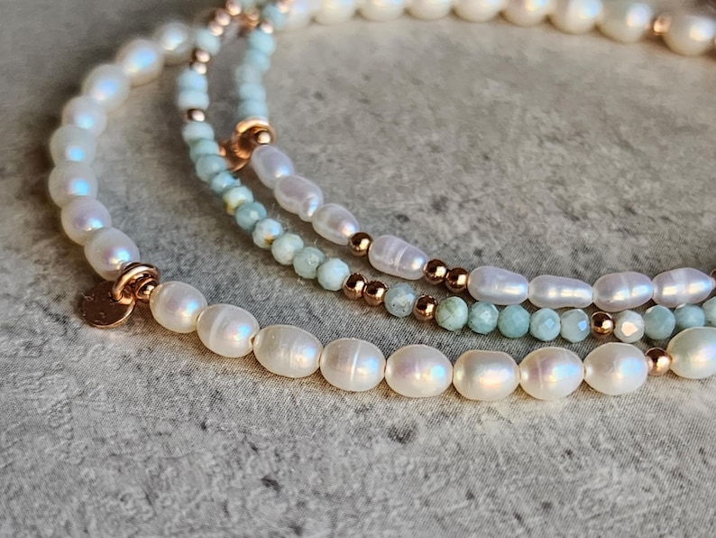 May include: Three layered bracelets with white oval pearls, small light blue beads, and rose gold accents. The bracelets are arranged in a curved pattern, showcasing the different textures and colors of the jewelry.