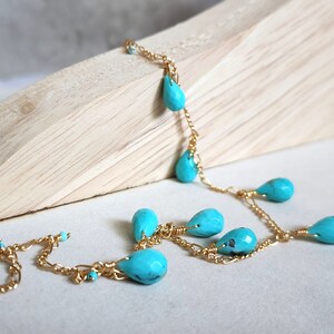 May include: A delicate gold chain necklace featuring teardrop-shaped turquoise beads. The necklace is displayed on a light-colored wooden surface, showcasing the vibrant blue of the gemstones. The necklace is a piece of jewelry.