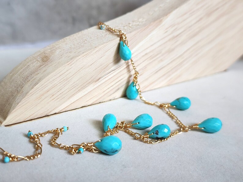 May include: A delicate gold chain necklace featuring teardrop-shaped turquoise beads. The necklace is displayed on a light-colored wooden surface, showcasing the vibrant blue of the gemstones. The necklace is a piece of jewelry.