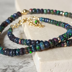 Op de afbeelding: Een set van een armband en ketting met gefacetteerde donkere opaal kralen met iriserende groene en blauwe flitsen, geaccentueerd met kleine goudkleurige metalen kralen, een goudkleurige karabijnsluiting en een verstelbare ketting met een opaal bedel.