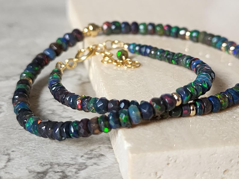 Op de afbeelding: Een set van een armband en ketting met gefacetteerde donkere opaal kralen met iriserende groene en blauwe flitsen, geaccentueerd met kleine goudkleurige metalen kralen, een goudkleurige karabijnsluiting en een verstelbare ketting met een opaal bedel.