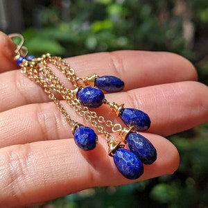 May include: Gold-tone earrings with multiple strands of delicate chain. Each strand features a faceted, deep blue gemstone. The earrings are held in a hand, showcasing the intricate design and color contrast against the skin.