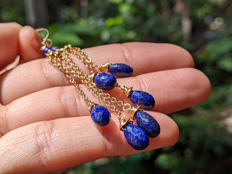 May include: Gold-tone earrings with multiple strands of delicate chain. Each strand features a faceted, deep blue gemstone. The earrings are held in a hand, showcasing the intricate design and color contrast against the skin.