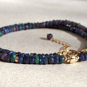 Op de afbeelding: Een ketting met kleine, gefacetteerde donkerblauwe en zwarte opaal kralen met iriserende groene en rode flitsen. De streng is geaccentueerd met kleine goudkleurige tussenstukjes en bevat een goudkleurige metalen sluiting met een verstelbare ketting.