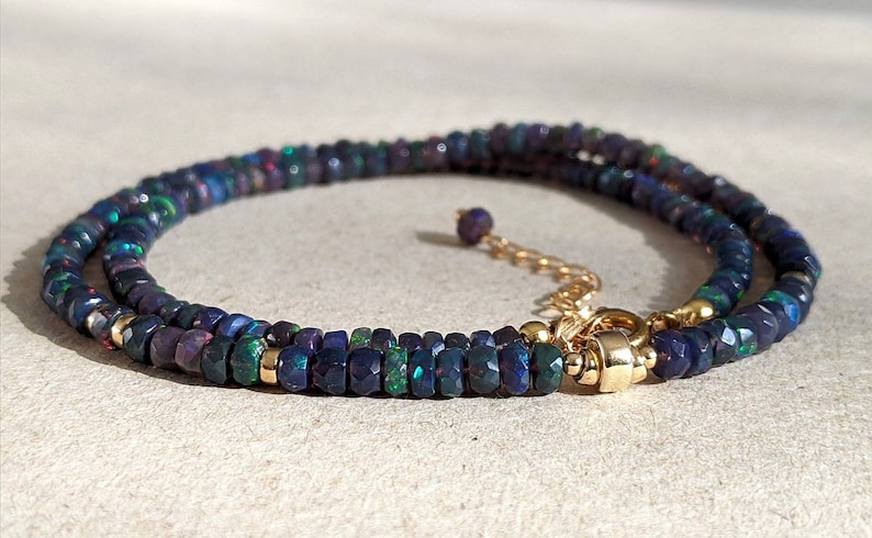 Op de afbeelding: Een ketting met kleine, gefacetteerde donkerblauwe en zwarte opaal kralen met iriserende groene en rode flitsen. De streng is geaccentueerd met kleine goudkleurige tussenstukjes en bevat een goudkleurige metalen sluiting met een verstelbare ketting.