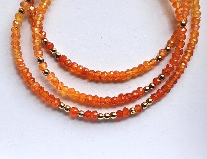 May include: Three strands of orange gemstone necklaces with gold-colored beads. The necklaces are made of small, faceted beads in shades of orange and yellow. The gold beads are evenly spaced along the strands, adding a touch of elegance.