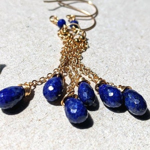 May include: Gold-tone earrings with dangling, faceted, deep blue lapis lazuli teardrop beads. The earrings feature delicate gold chains and small gold beads, creating a layered, elegant design. The earrings are displayed on a neutral surface.