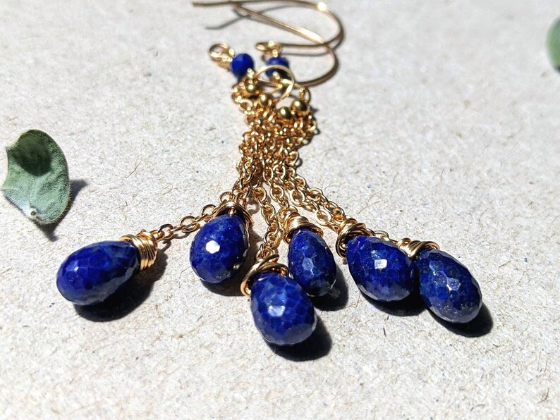 May include: Gold-tone earrings with dangling, faceted, deep blue lapis lazuli teardrop beads. The earrings feature delicate gold chains and small gold beads, creating a layered, elegant design. The earrings are displayed on a neutral surface.