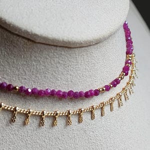 Pu&ograve; includere: Primo piano di due collane. La collana superiore presenta piccole perline sfaccettate di colore rosa intenso. La collana inferiore &egrave; dorata con un design a frange. Entrambe le collane sono esposte su una superficie di colore neutro.