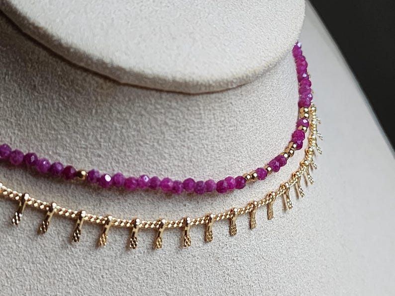 Pu&ograve; includere: Primo piano di due collane. La collana superiore presenta piccole perline sfaccettate di colore rosa intenso. La collana inferiore &egrave; dorata con un design a frange. Entrambe le collane sono esposte su una superficie di colore neutro.