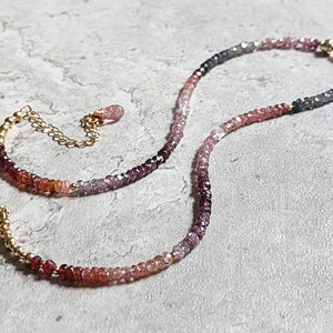 May include: A delicate necklace with a gradient of small, colourful gemstones. The stones transition from deep reds to pinks, purples, and greys. The necklace has a gold chain and clasp, with a small dangling charm.