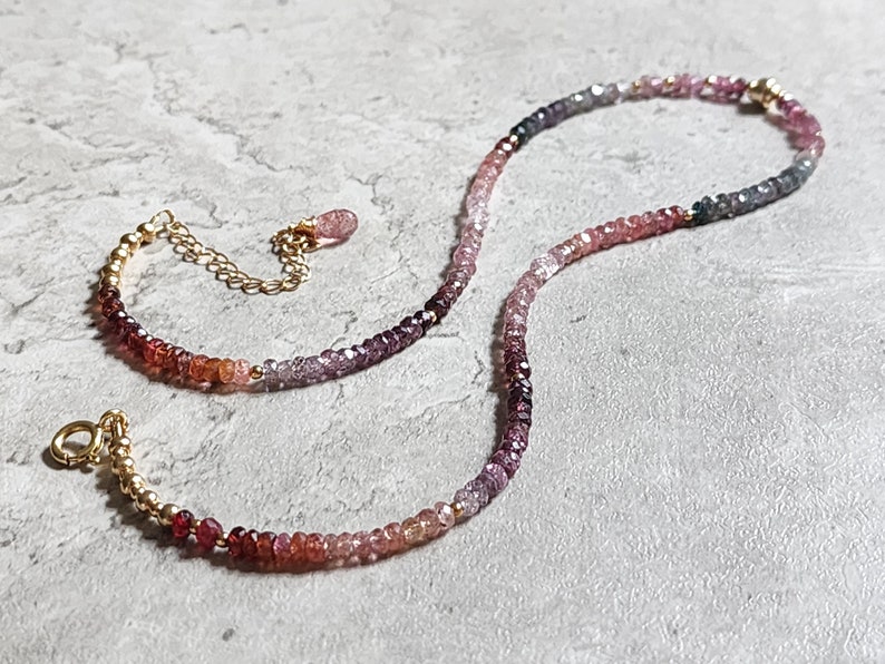May include: A delicate necklace with a gradient of small, colourful gemstones. The stones transition from deep reds to pinks, purples, and greys. The necklace has a gold chain and clasp, with a small dangling charm.