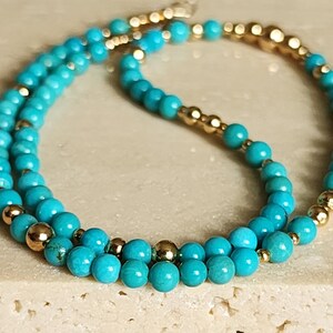 May include: A turquoise and gold beaded necklace. The necklace is made of small, round turquoise beads and gold beads. The turquoise beads are arranged in a double strand, with the gold beads interspersed between the turquoise beads.