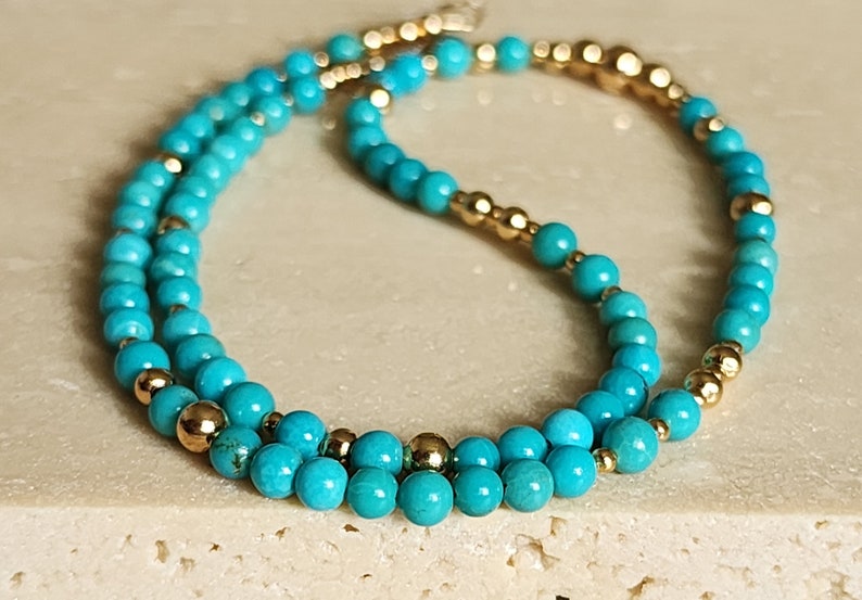 May include: A turquoise and gold beaded necklace. The necklace is made of small, round turquoise beads and gold beads. The turquoise beads are arranged in a double strand, with the gold beads interspersed between the turquoise beads.